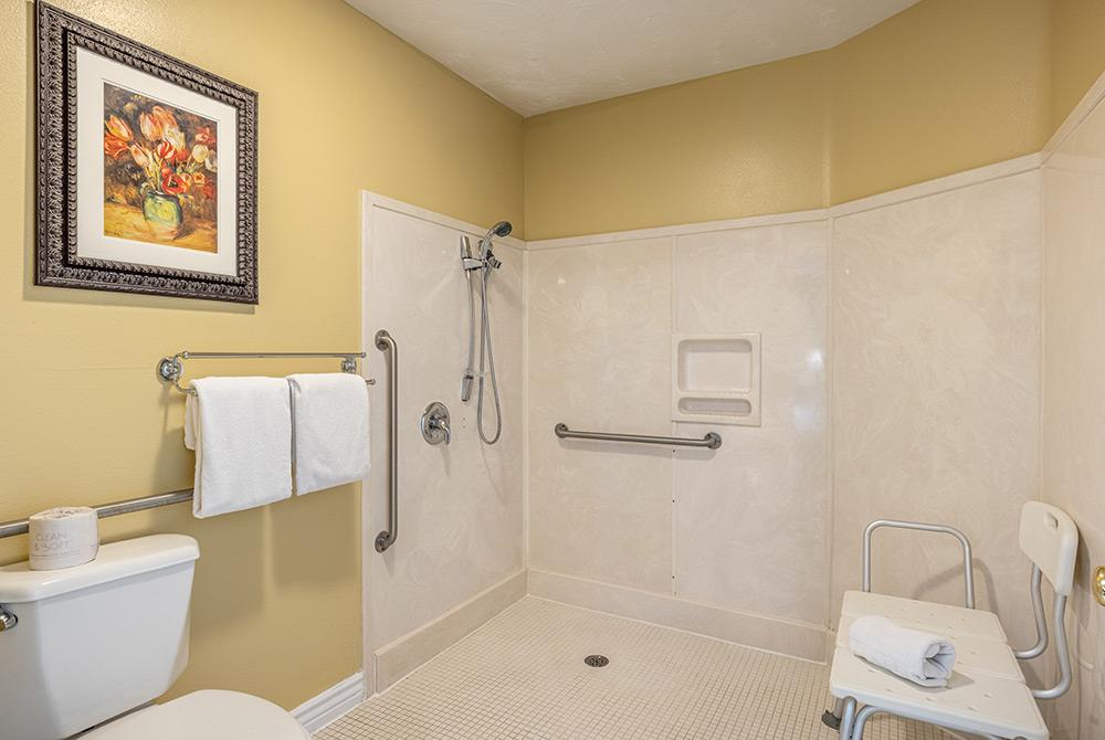 Accessible bathroom in our accessible king room Kanab UT, featuring a roll-in shower, grab bars, handheld showerhead, a shower chair, towels on racks, and a toilet with toilet paper. Framed floral artwork hangs on the yellow wall.