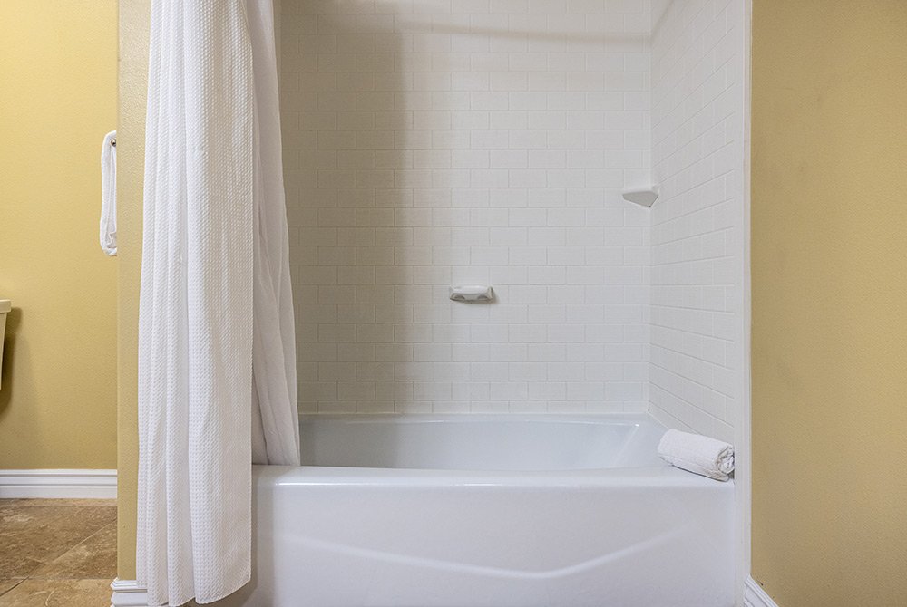 A white bathtub with a white tile shower wall, a white shower curtain partially pulled back, and a rolled white towel on the tub edge, in a bathroom with beige walls—perfectly appointed for your stay in our standard king room in Kanab, UT.