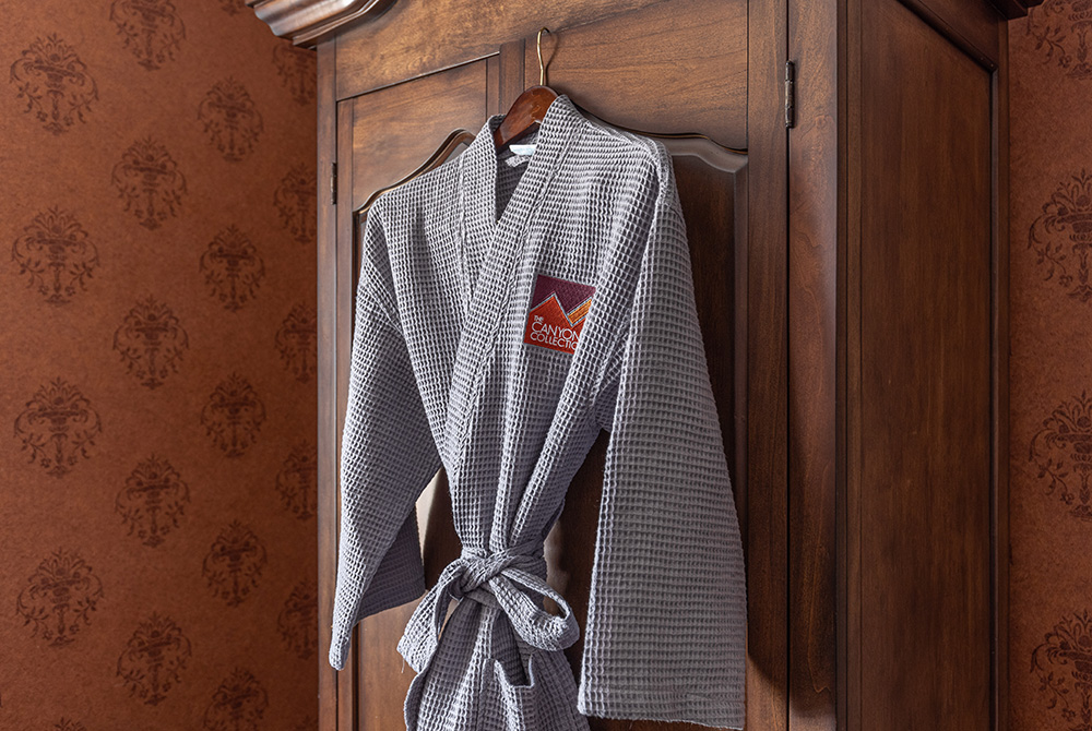 A gray waffle-textured bathrobe with a logo hangs on a wooden wardrobe against a patterned brown wallpaper background, adding an elegant touch to the deluxe double queen room in Kanab, UT.