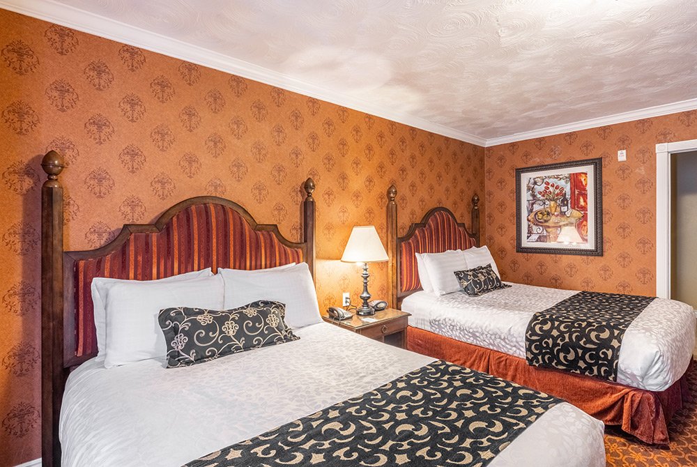 Experience the comfort of a deluxe double queen room Kanab UT, featuring two queen beds with decorative headboards and patterned bedding in a warmly lit hotel setting. A lamp sits on a nightstand between the beds, while framed artwork adorns the textured, orange-patterned wallpapered wall.
