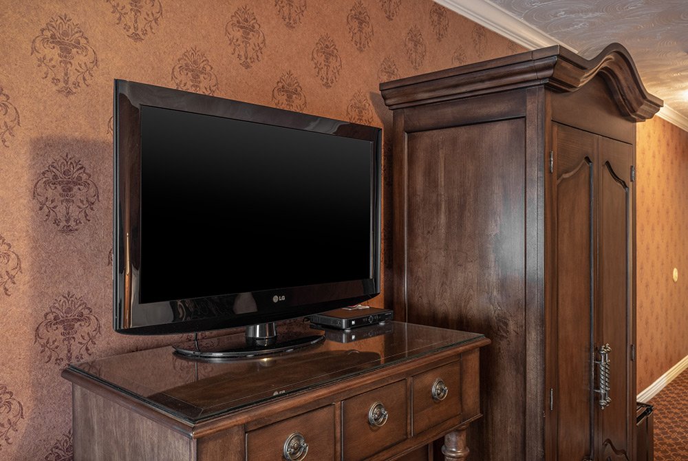 A flat-screen TV sits on a wooden dresser next to a tall wooden wardrobe in a deluxe double queen room Kanab UT, featuring patterned wallpaper and carpet.