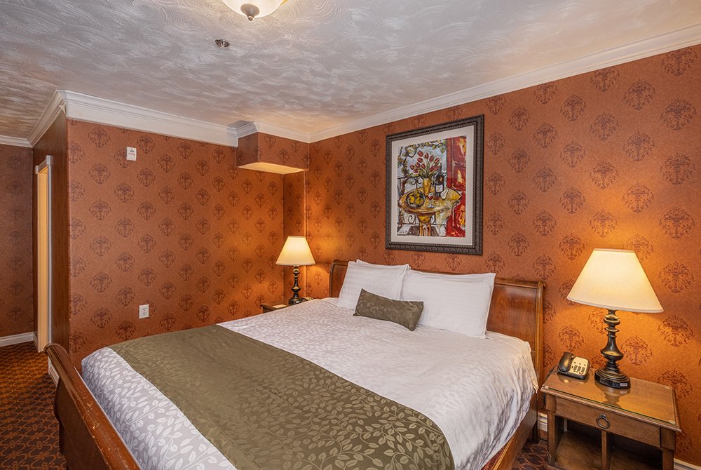 Enjoy a cozy standard king room Kanab UT featuring a king bed, white and olive bedding, two lamps, wooden nightstands, patterned wallpaper, and a framed painting above the headboard.