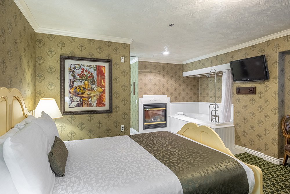 Hotel room with a neatly made bed, patterned green bedding, wall art, a fireplace, a TV mounted on the wall, and a white jacuzzi tub in the corner. The walls have green floral wallpaper.