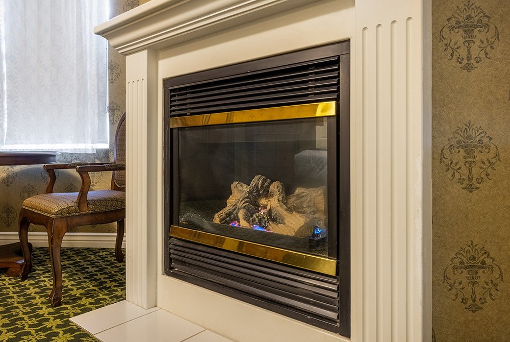 A gas fireplace with decorative logs is set in a cream-colored mantel, adding warmth and elegance to this deluxe king room in Kanab, UT. To the left, a wooden chair with an upholstered seat sits on a green patterned carpet near a window with sheer curtains.