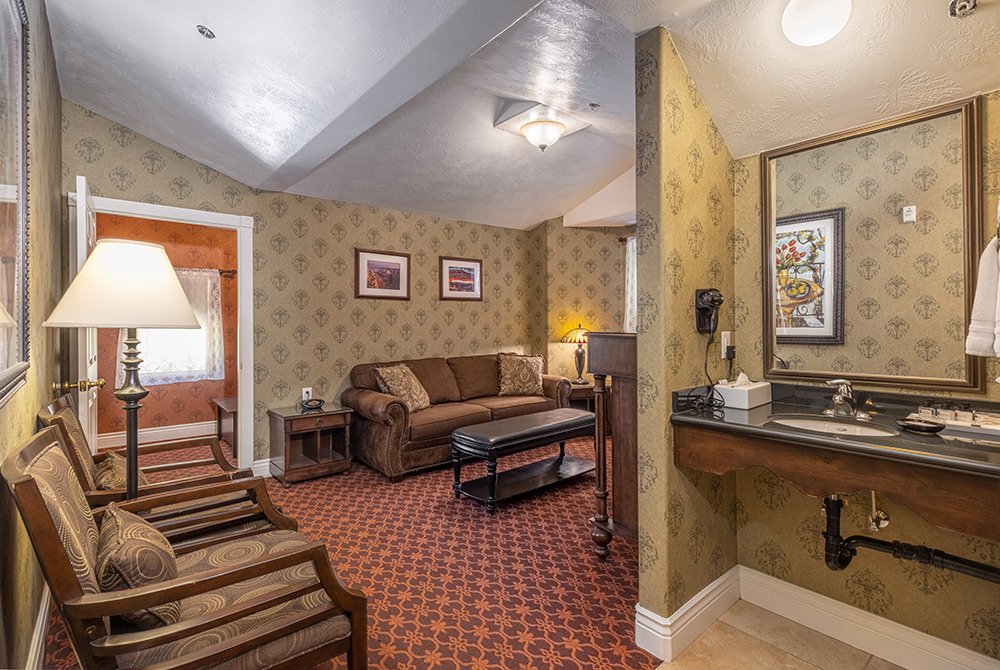 Cozy 2 bedroom suite in Kanab UT with patterned walls and carpet, a brown sofa, two chairs, side tables, lamps, framed art, and an open vanity area with a sink and mirror. Doorway leads to another warmly lit room.