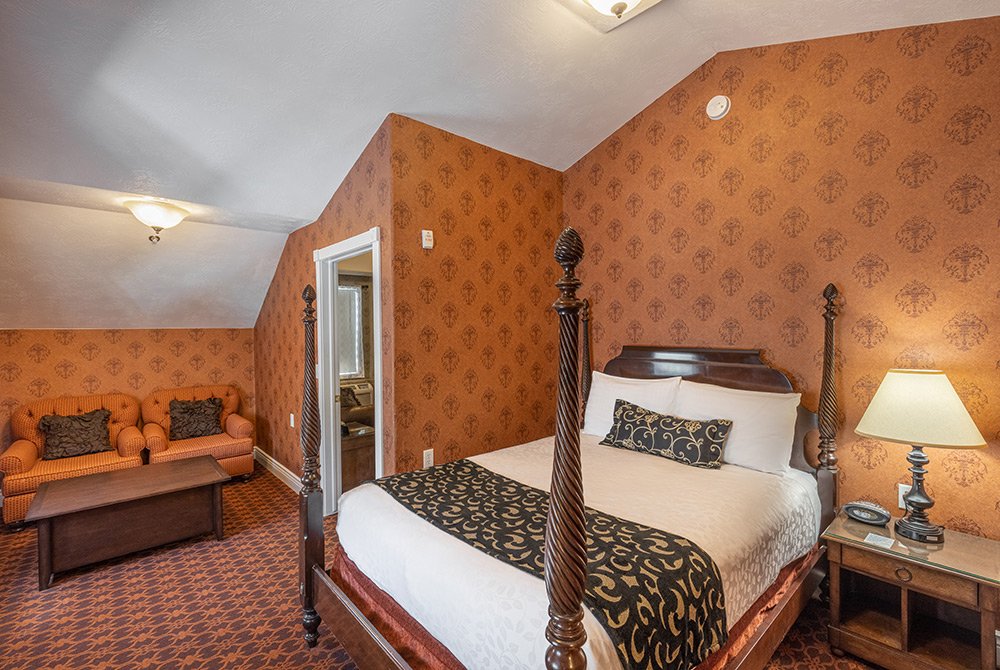 A cozy 2 bedroom suite in Kanab, UT features a charming bedroom with a four-poster bed, patterned brown wallpaper, matching carpet, a wooden nightstand with a lamp, and an orange sofa with a coffee table in the corner.