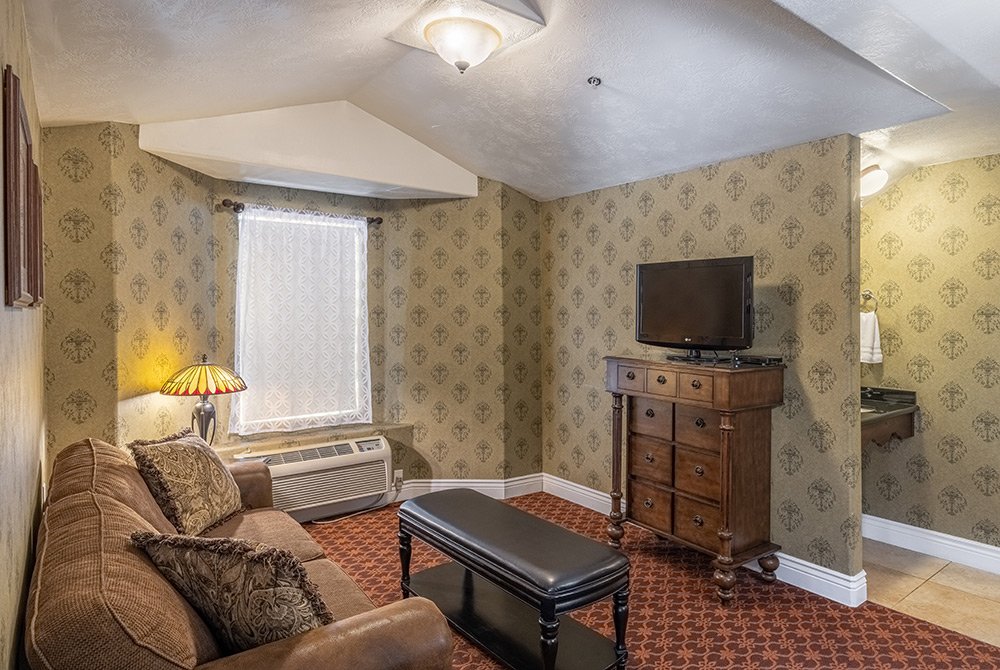A cozy living room in a 2 bedroom suite in Kanab, UT, featuring a brown couch, patterned carpet, small table, and a dresser with TV. The space is adorned with vintage wallpaper and a lamp sits by the sofa beneath a window draped with lace curtains. A bathroom is partly visible from this inviting setting.