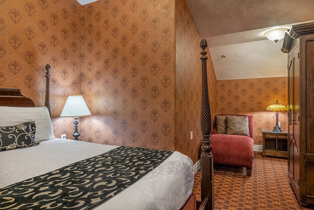 A cozy bedroom within a 2 bedroom suite in Kanab UT features patterned wallpaper, a canopy bed, ornate lamps, a red loveseat, and a wooden wardrobe. Warm lighting and detailed decor create an inviting, vintage atmosphere.