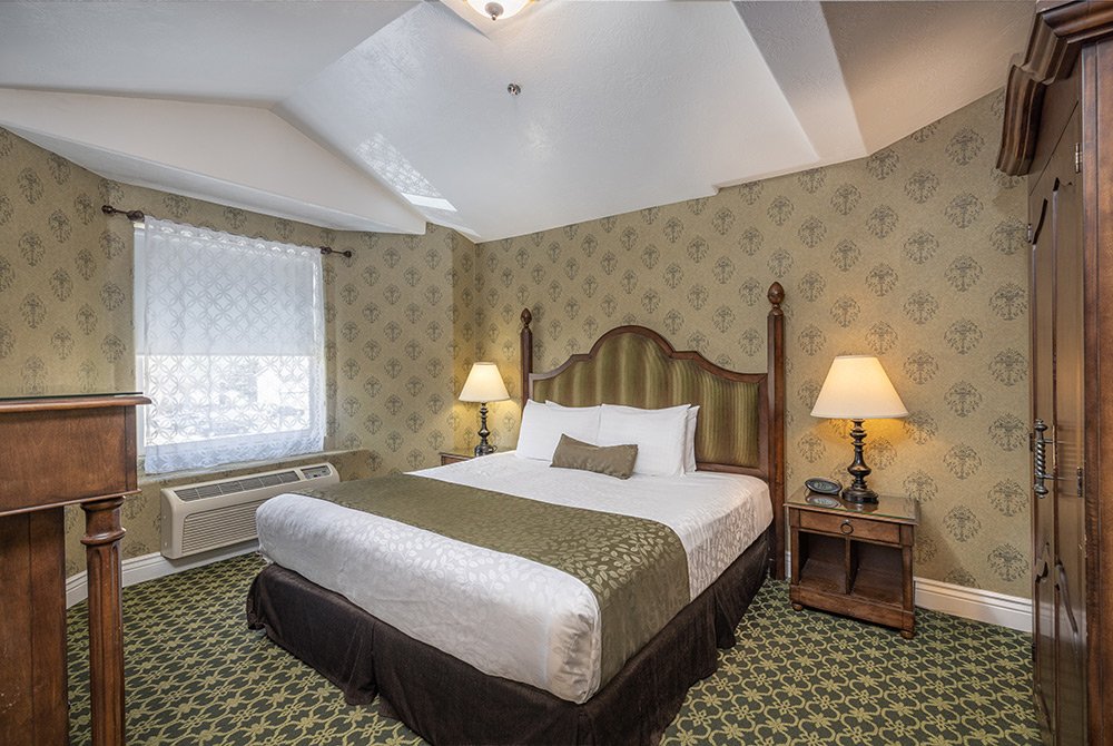 A cozy 2 bedroom suite in Kanab, UT, featuring a spacious bedroom with a large bed, two bedside tables with lamps, patterned wallpaper, a window with lace curtains, and a green carpet adorned with a charming floral design.