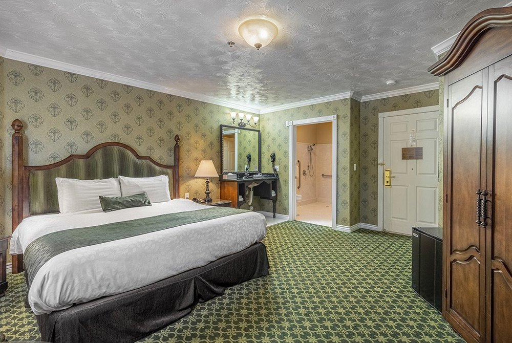 Experience the charm of an accessible king room in Kanab, UT, featuring a large bed, green patterned carpet, classic wooden furniture, a wardrobe, and a desk with a mirror. An open doorway leads to the bathroom, while vintage-style wallpaper and lighting add timeless character to your stay.