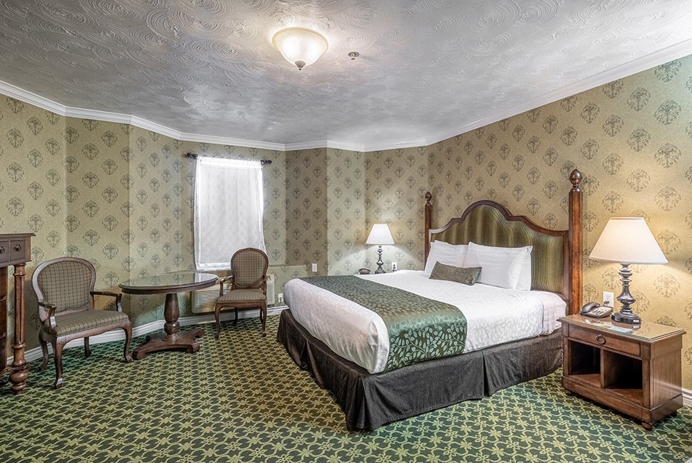 Experience timeless elegance in our accessible king room Kanab UT. This inviting hotel room features a king bed, green patterned carpet and wallpaper, two chairs at a round table, two lamps, and a window with light curtains. With charming vintage decor and thoughtful accessibility features, your stay in Kanab will be as comfortable as it is stylish.